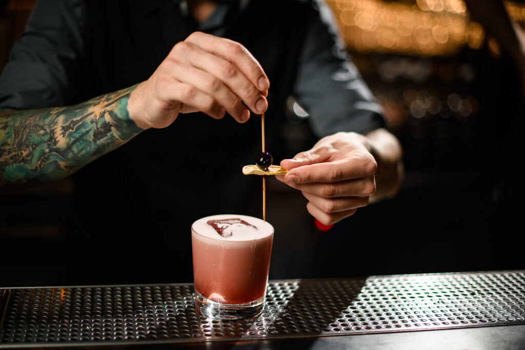 Close-up of tattooed male bartender adding fresh olive to alcohol drink