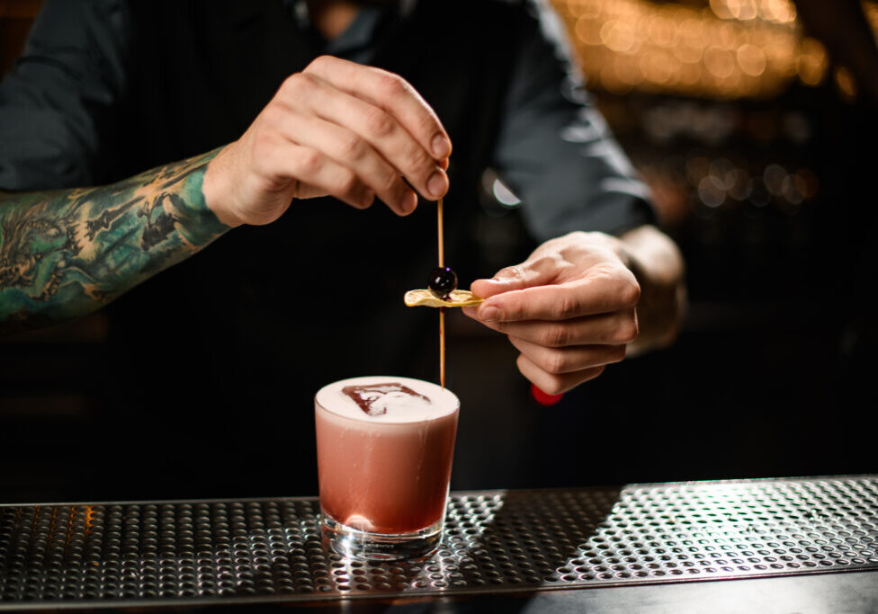 Close-up of tattooed male bartender adding fresh olive to alcohol drink